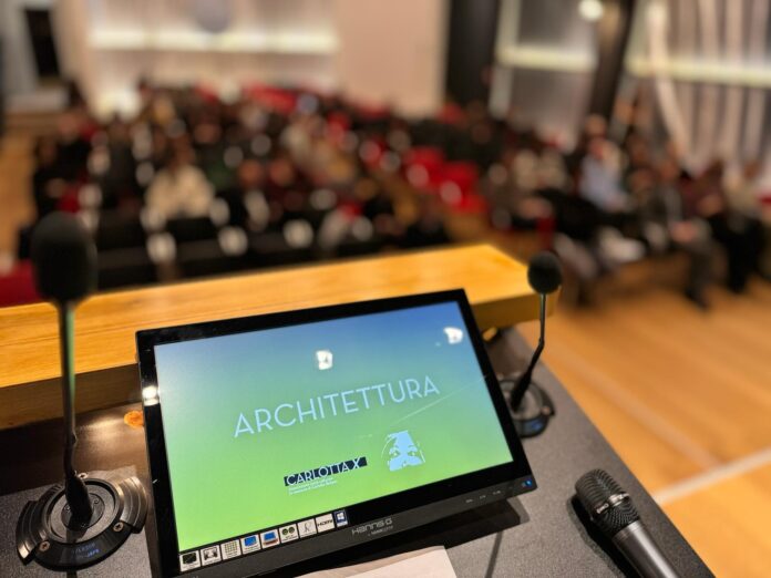 DEsign - uno schermo di pc con accanto un microfono, in un auditorium con tant agente seduta