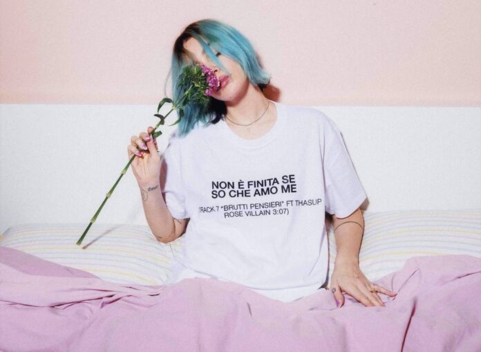 rose villain seduta a letto, indossa una t shirt bianca e ha una rosa nella mano destra