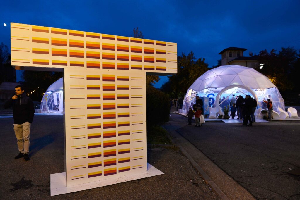 Biennale Tecnologia - Una grande lettera "T" illuminata di arancione nel crepuscolo della sera, la lettera è in mezzo a una piazza dove si vedono delle persone che entrano ed escono da una struttora rotonda e bianca a forma di cupola