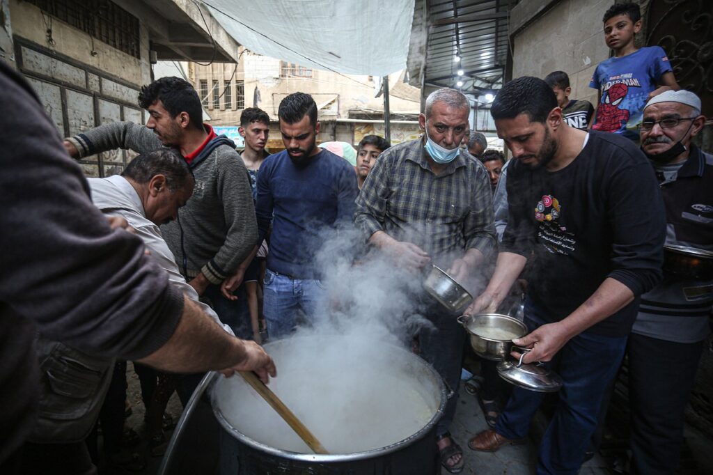 Gaza, delle persone stanno in coda per prendere del cibo, in primo piano un enorme pentolone con del cibo fumante