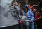 Emergenza fame a Gaza . un uomo verso del cibo da un pentolino in una pentola più grossa sorretta da un bambino, Lo scenario è di guerra, intorno macerie e persone in fila