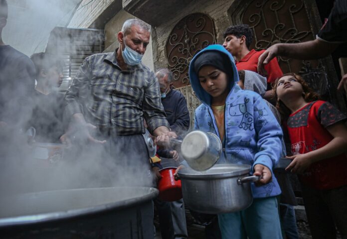 Emergenza fame a Gaza . un uomo verso del cibo da un pentolino in una pentola più grossa sorretta da un bambino, Lo scenario è di guerra, intorno macerie e persone in fila