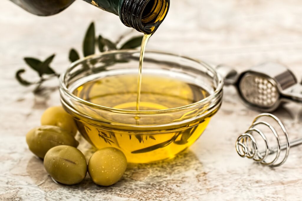 Una coppetta di vetro trasparente con dell'olio d'oliva. Vicino delle olive verdi e dietro un ramoscello di ulivo