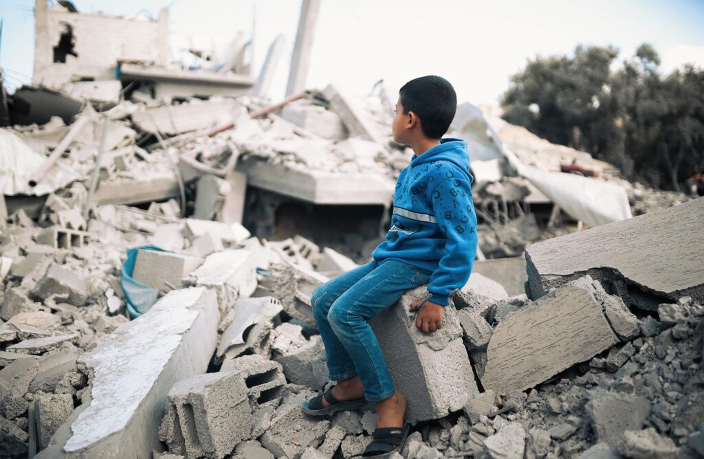 Emergenza fame Gaza - un bambino è seduto con una tuta blu, sul cumulo di macerie di una casa distrutta dai bombardamenti a Gaza