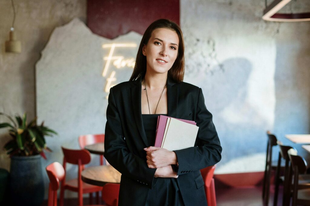 lavoro giovani - una ragazza in primo piano vestita elegante in nero, tiene in mano una cartellina di fogli, dietro di lei dei tavoli e delle sedie