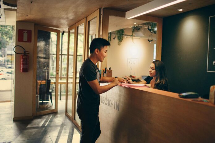 Lavoro per i giovani - nella foto la reception di un hotel con una ragazza dietro il bancone che sta compilando i dati del cliente giovane che sta davanti al bancone. L'amibente è con luci soffuse e si vede una porta vetro da cui si intravede un giardino