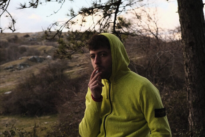 samuele cara fotografato all'aperto, indossa una felpa gialla con cappuccio, è intento a fumare una sigaretta