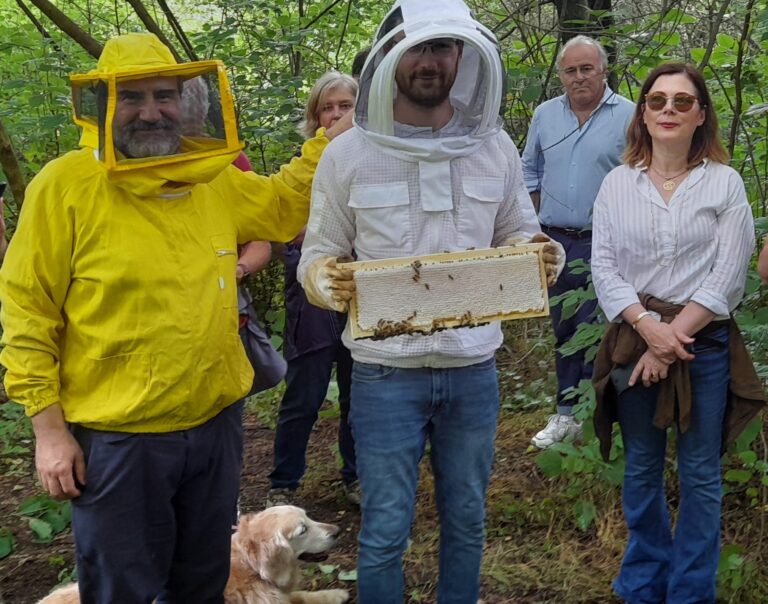 orti e fiori - un gruppo di persone. In primo piano due uomini indossano dei caschi protettivi per le api, uno di colore giallo, l'altro biacno. Di fianco una donna in camicetta e jeans 