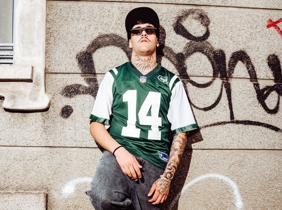 le-one indossa una maglietta bianco-verde d baseball, cppellino e occhiali da sole. E' appoggiato a un muro con dei graffiti