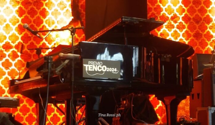 Serata finale Premio Tenco 2024 - un pianoforte a coda nero con la scritta Tenco 2024, sul palco di un teatro illuminato da luci arancioni