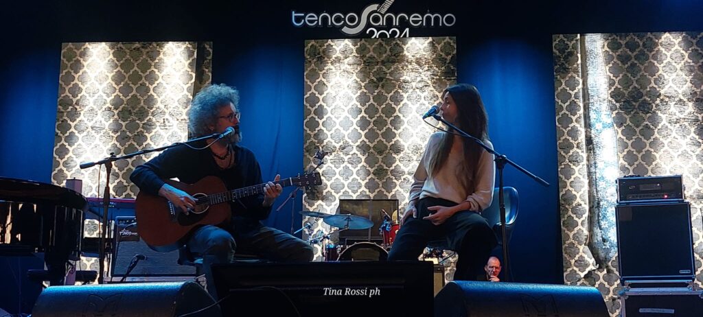Serata finale Premio Tenco 2024 - Simone Cristicchi a sinistra con folti capelli ricci, occhiali, suona la chitarra, a destra Amara, con lunghi capelli neri