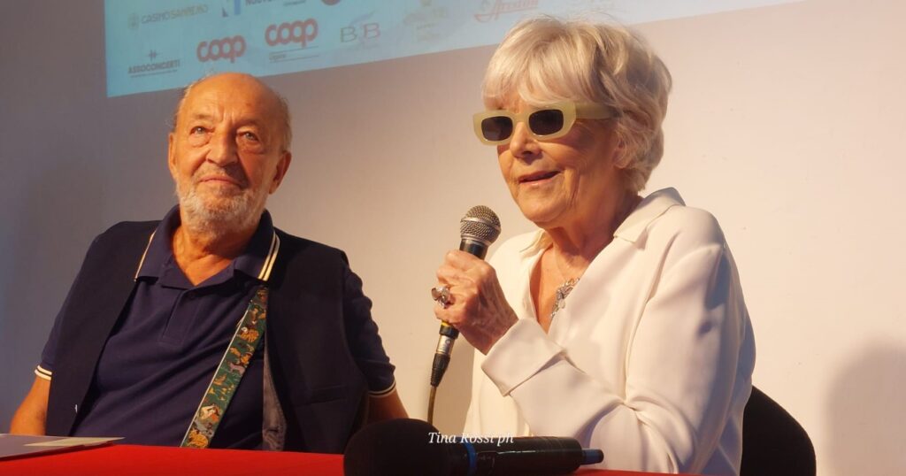 Caterina Caselli, vapelli biondi, occhiali da sole neri, maglia bianca parla al microfono in conferenza stampa, vicino a lei un uomo con maglia nera