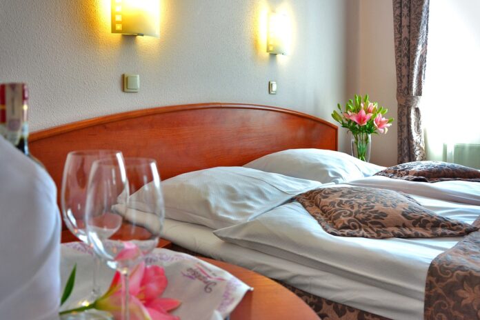 una camera d'albergo di cui si vede il letto con lenzuola bianche e in primo piano un vassoio con due flutes e dei fiori rosa