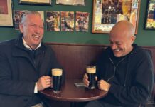 Max Pezzali e Mauro Repetto seduti a un tavolino del bar con due birre scure ridono divertiti
