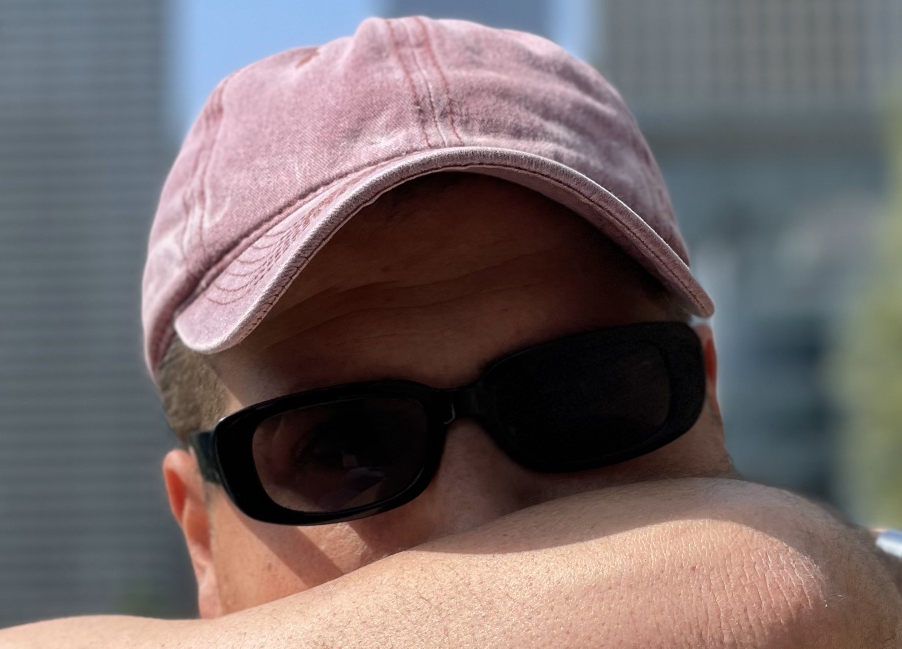 estro large in primo piano, il volto parzialmente coperto da un avambraccio, indossa cappellino da baseball e occhiali scuri