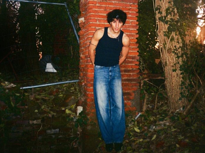 still charles, appoggiato a una colonna di mattoni, in mezzo al verde, indossa jeans blu e canotta nera