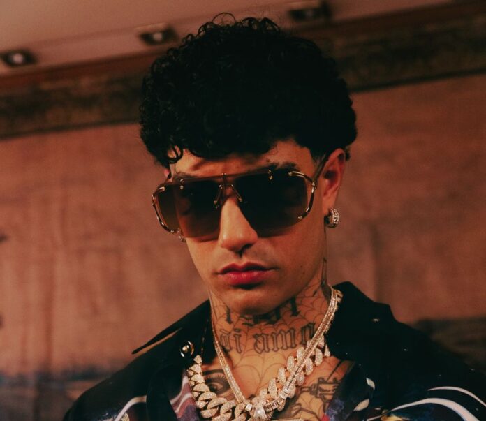Tony Effe a Sanremo - primo piano del rapper con capelli ricci corti e ciuffo sulla fronte, occhiali da sole scuri e tante collane al collo, espressione seria