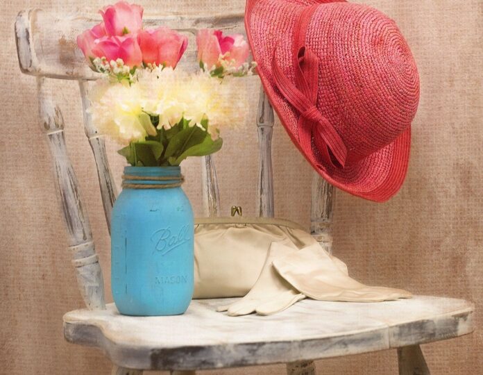 arredamento sostenibile e mobili usati - una sedia di legno vecchia ridipinta di bianco con sopra dei guanti di raso da signora appoggiati a una pochette avorio, un vaso di fiornri azzurro e un cappello rosso con dei fiori rossi appoggiato allo schienale