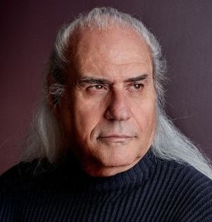 franco mussida in primo piano, ha i capelli lunghi e bianchi e indossa una maglia nera