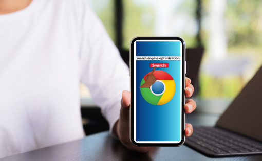 Google Chrome - nella foto una persona con t shirt bianca è seduta a un ascrivania, è sfocata, tiene in mano e mostra la schermata di un cellulare che figura uno sfondo azzurro con un cerchio colorato di verde, blu, giallo e rosso con la scritta 