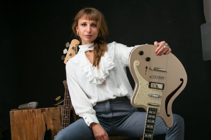 Gloria Abbondi in primo piano, indossa jeans e camicia bianca e ha in mano una chitarra