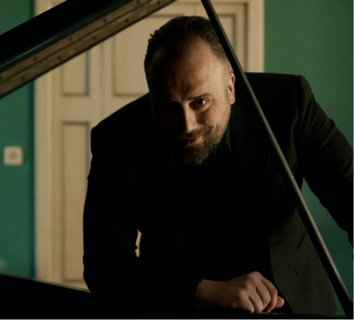 raphael gualazzi in primo piano, vestito di scuro, appoggiato a un pianoforte a coda