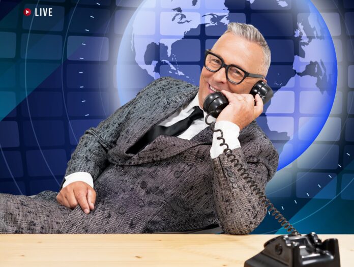 paolo belli, indossa un vestito grigio, camicia bianca e cravatta nera. è appoggiato a una scrivani, intento a parlare al telefono