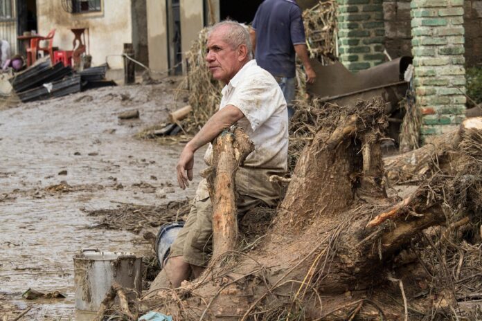 Ravenna alluvione Romagna - nella foto un uomo anziano in maniche corte è seduto per terra tra il fango e i detriti di un 'alluvione e appoggia il braccio su un tronco d'albero portato dal fango