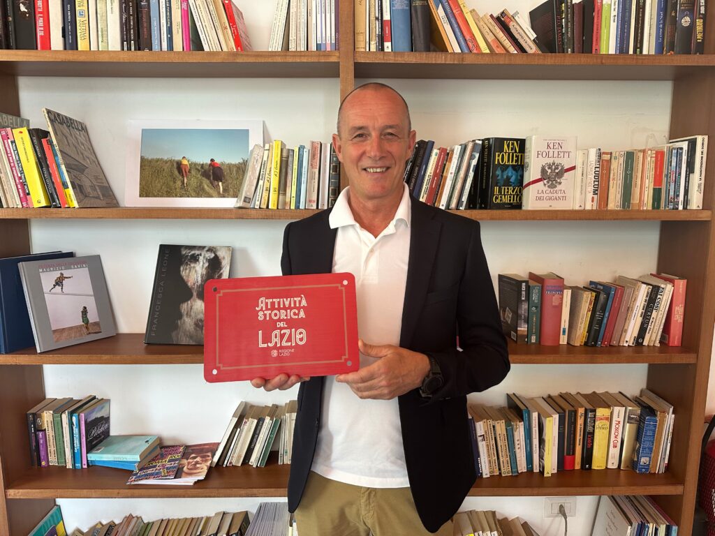 Sperlonga Antico Caffè Trani - il proprietario del locale veste una giacca nera con sotto una polo bianca, tiene in mano la targa di riconoscimento, è rossa, dietro di lui una libreria piena di libri 