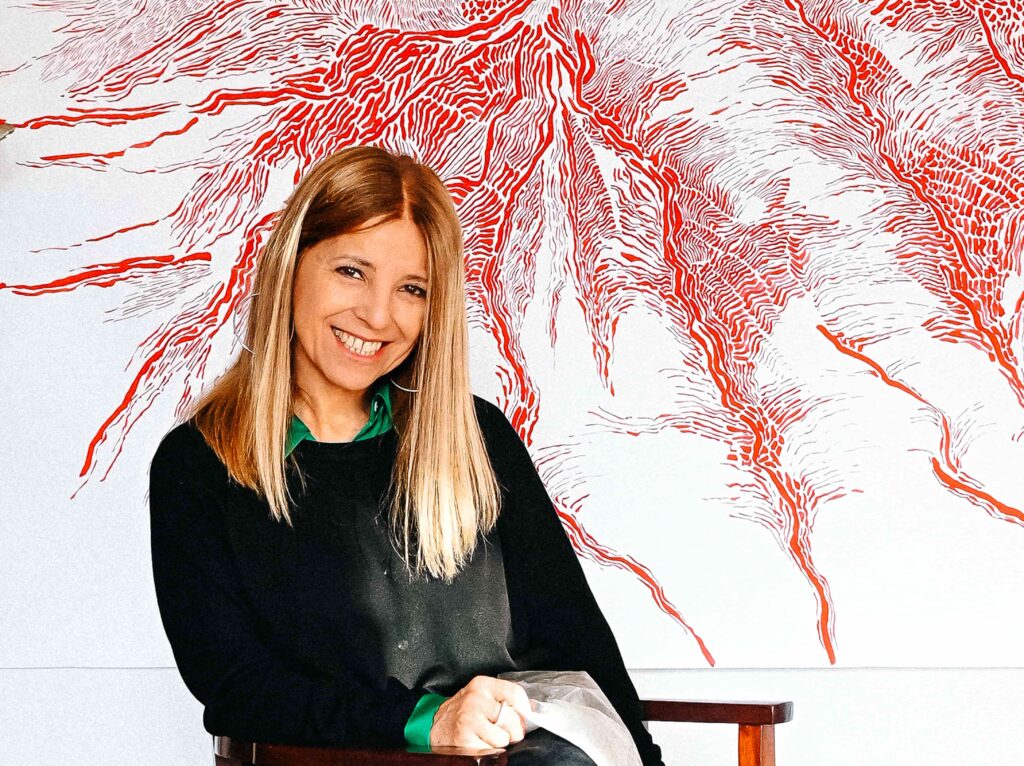 Art Site Fest 2025 a Moasca arte, Paesaggi Siderali e vino tra le colline del Monferrato - nella foto l'artista seduta davanti a una sua opera , è bionda, giovane, sorride, indossa una giacca nera su camicetta verde
