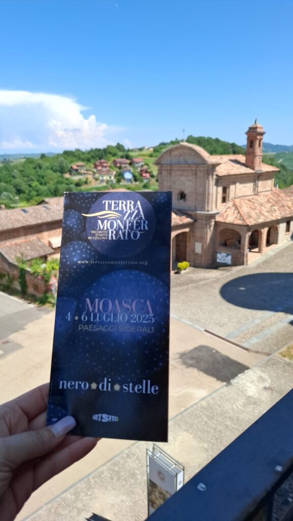 Una veduta del Castello di Moasca (sullo sfondo) e in primo piano una mano tiene la brochure dell'evento