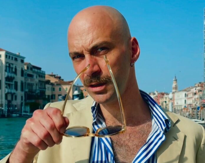 azzurrissimo - amalfitano in primo piano indossa giacca beige e camicia a righe, ha in mano gli occhiali da sole