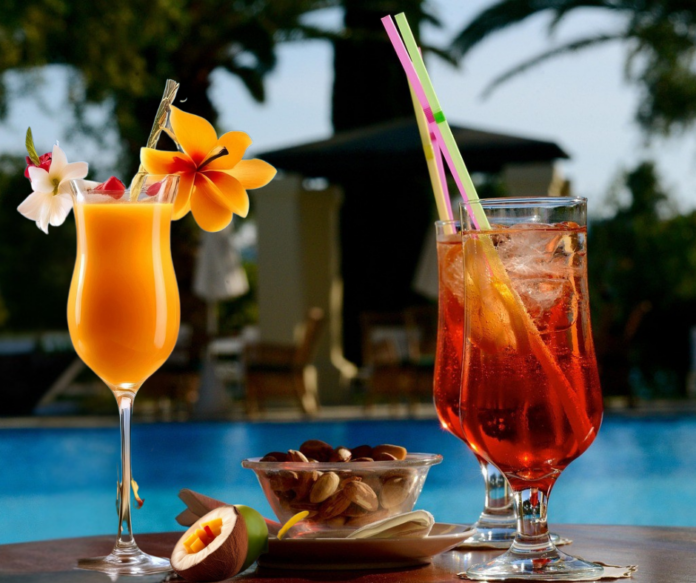 Cocktail - nella foto tre bicchieri fatti a campana, a sinistra uno è pieno di una bevanda color pesca e ha una cannuccia e dei fiori di guarnizione, a destra, due bicchieri con un abevanda rossa, con cannuccia. I bicchieri sono su un tavolo di legno con una ciotola di noccioline e sullo sfondo il mare e una capanna di paglia