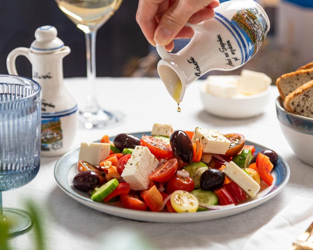Insalate estive creative l'estate si mangia a colori - nella foto una ciotola bianca con pomodoro formaggio bianco olive nere e una mano versa olio da un'ampolla