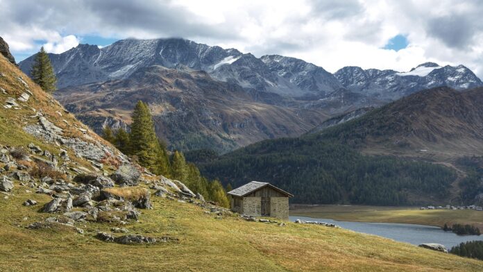 UNCEM il silenzio della montagna è un grido di aiuto - nella foto il versante di un amontagna con rocce e prato, una casetta isolata, a fianco scorre un fiume e sullo sfondo una catena di monti