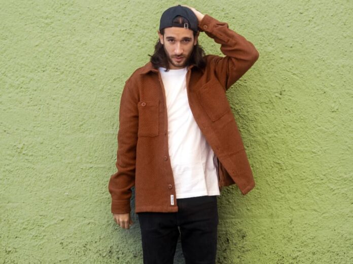 cappie in primo piano, indossa una t-shirt bianca, pantaloni neri e giacca rossa