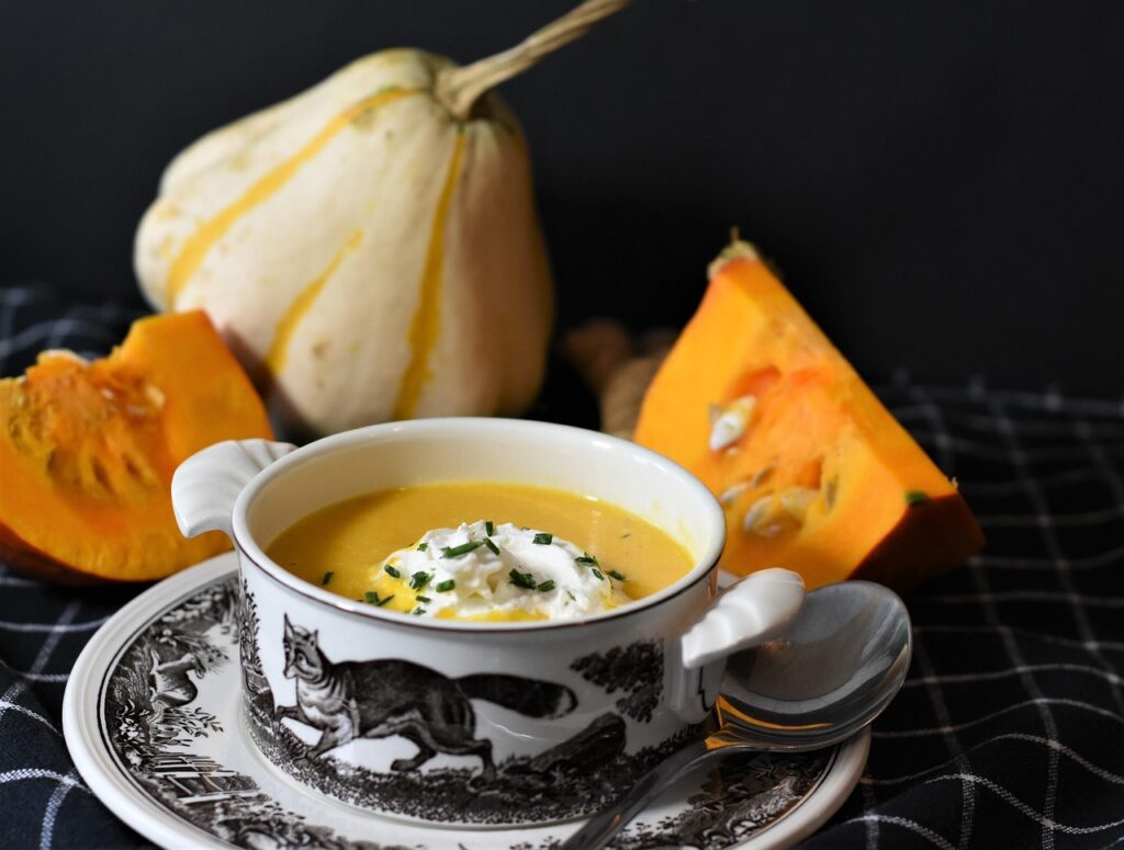 Ottobre ricette - una scodella da brodo con un azuppa di zucca con crema bianca, vicino alla tazza deipezzi di zucca gialla e una piccola zucca bianca