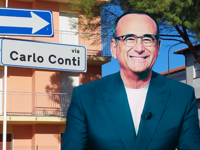 Chi sono i big in gara Sanremo 2026 - nella foto Carlo Conti in primo piano indossa una giacca blu sopra un amaglia bianca, occhiali da vista, sorride. Di fianco un cartello stradale con la scritta 