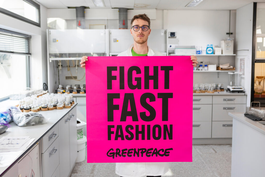 Abiti Shein - un uomo in un laboratorio chimico, con capelli neeri, occhiali da vista con montatura nera, tiene in mano un cartello rosa con la scritta "Fight Fast FAshion Greenpeace