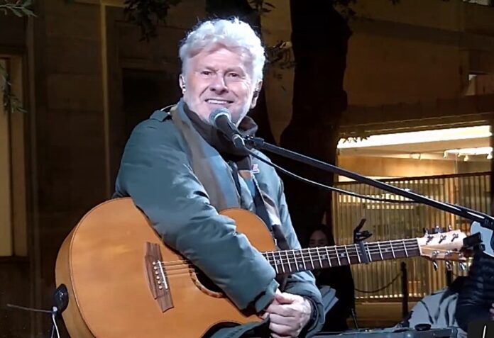 SLA LA PROMESSA 2025 - nella foto Ron con capelli ricci bianchi, vestito con giaccone verde, abbraccia la chitarra seduto su uno sgabello e sorride all'obbiettivo