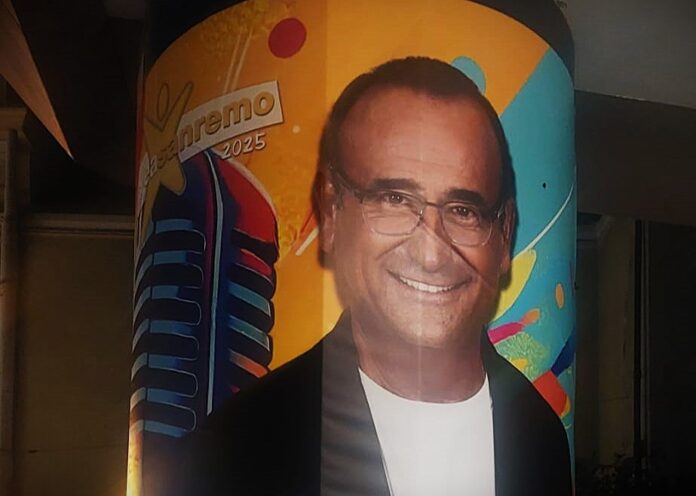 Area Sanremo - Zetatielle Magazine - foto di Tina Rossi Ph area sanremo - Area Sanremo un sogno o il grande inganno degli emergenti - Nella foto un pilastro con il poster di Carlo Conti con braccia incrociate, indossa giacca nera maglietta bianca, occhiali da vista, dietro di lui disegnato un microfono