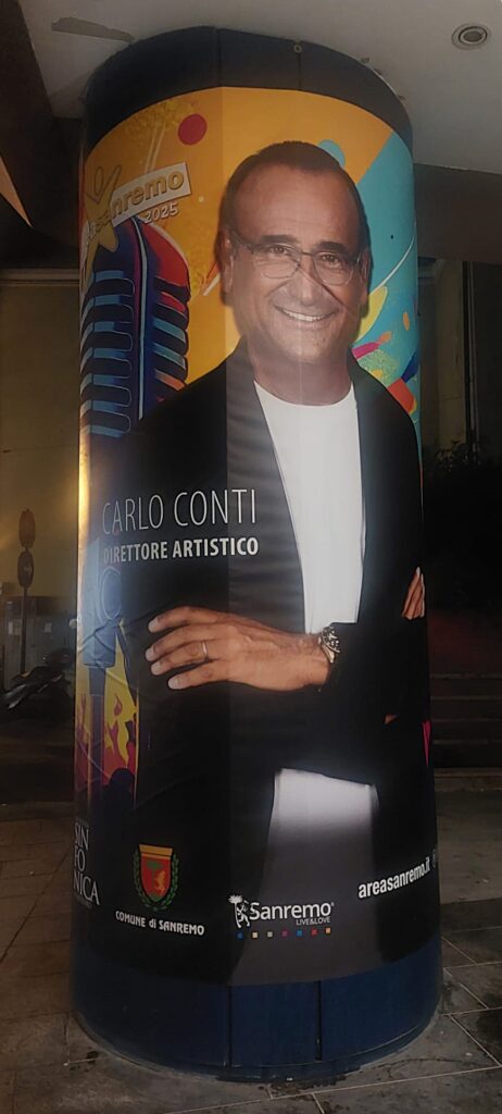 Area Sanremo un sogno o il grande inganno degli emergenti - Nella foto un pilastro con il poster di Carlo Conti con braccia incrociate, indossa giacca nera maglietta bianca, occhiali da vista, dietro di lui disegnato un microfono