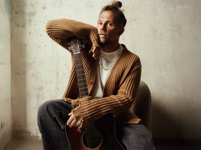 enrico nigiotti in primo piano, indossa jeans scuri, cardigan marrone e tiene tra le gambe una chitarra acustica