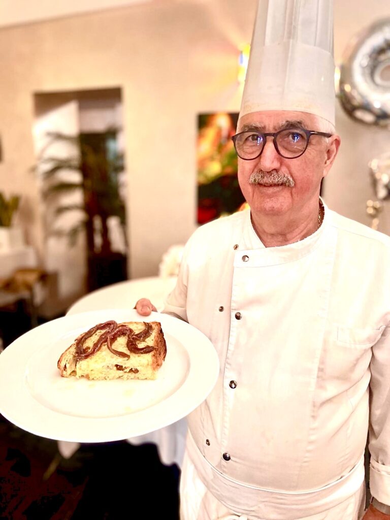 Nazareno Fontana vestito con divisa da chef bianca, cappello da cuoco bianco, tiene in mano un piatto bianco con sora un afetta di panettone con salsa di acciughe