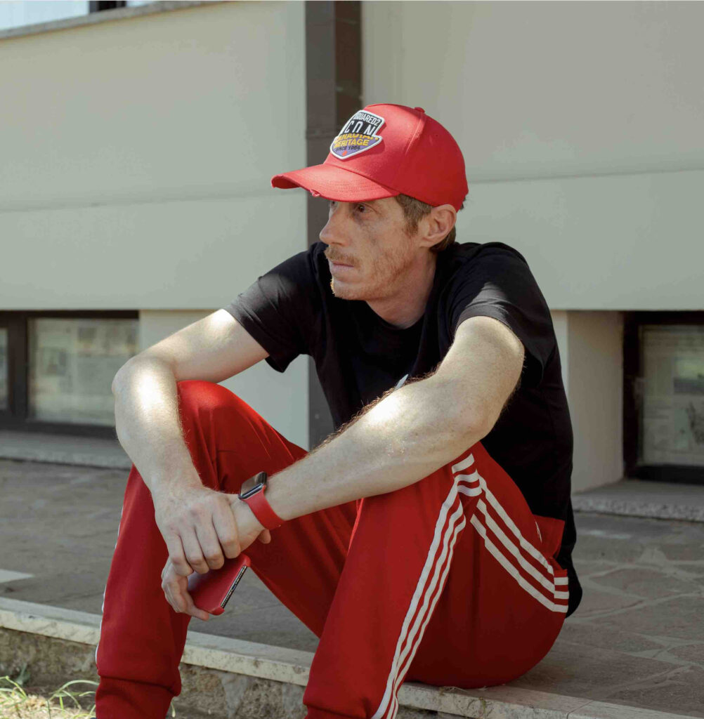 Luca Red seduto sul bordo di un marciapiede indossa cappellino con visiera rosso, t shirt nera, pantalone della tuta rosso con strisce bianche sul bordo
