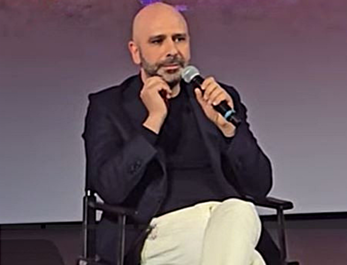 film di checco zalone - l'attore in primo piano, indossa pantaloni chiari, t-shirt e giacca nera, e tiene il microfono nella mano sinistra