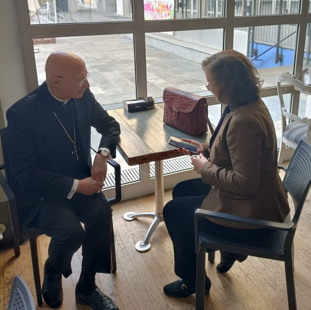 Maura Maffei intervista il cardinale. Entrambi sono seduti a un tavolo davanti a una vetrata.