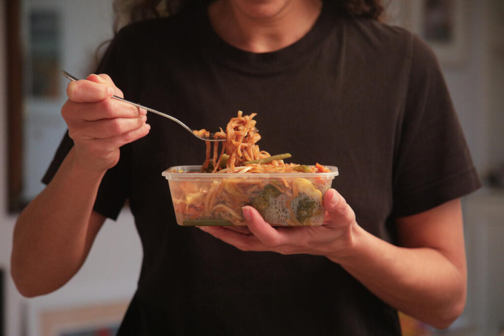 cibi pronti - nella ofto si vede una persona con t shirt nera che sta mangiando con una forchetta del cibo che è in una vaschetta di plastica