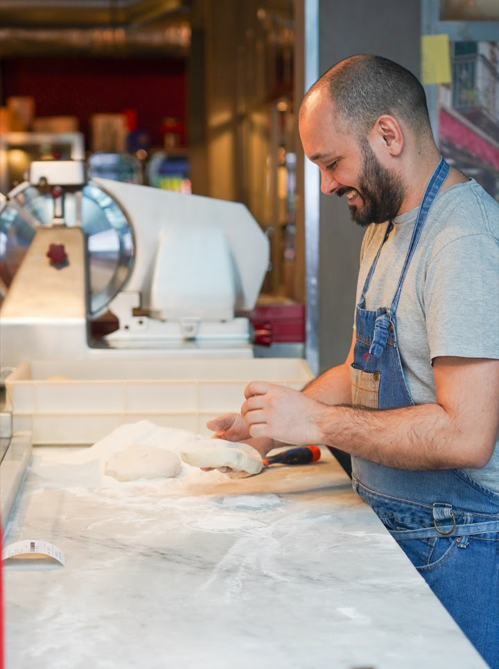Corso professionale disoccupati pizzaioli - un uomo di profilo è dietro un bancone da lavoro di una pizzeria e sta preparando un apizza. Sul tavolo c'è tanta farina e degli impasti