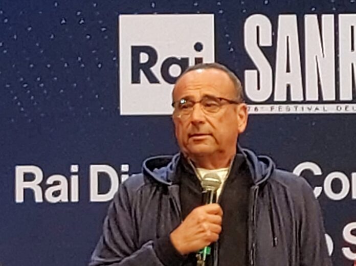 la scaletta prima serata - carlo conti veste una tuta blu con microfono in mano e indossa occhiali